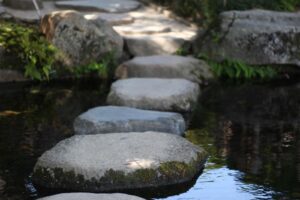 Vier gro&szlig;e grob behauene Steine f&uuml;hren &uuml;ber einen Fluss ans helle Ufer.
